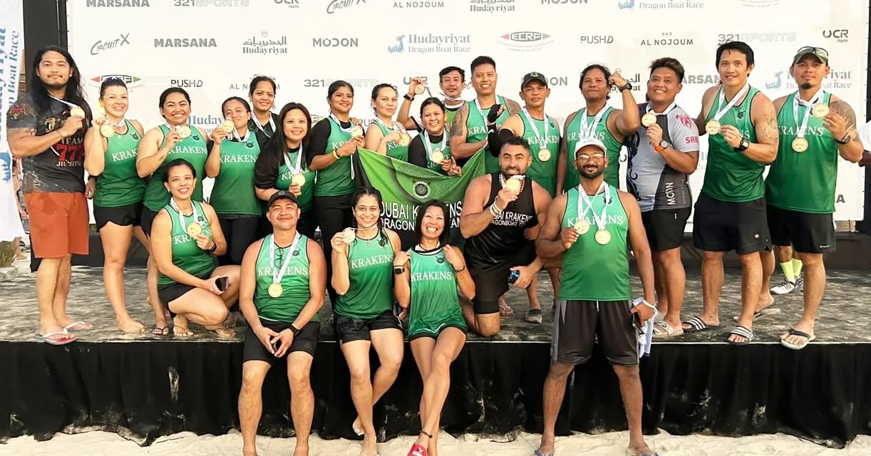 The image depicts a diverse group of 19 individuals, predominantly adults, gathered for a photograph on a stage with a bla...